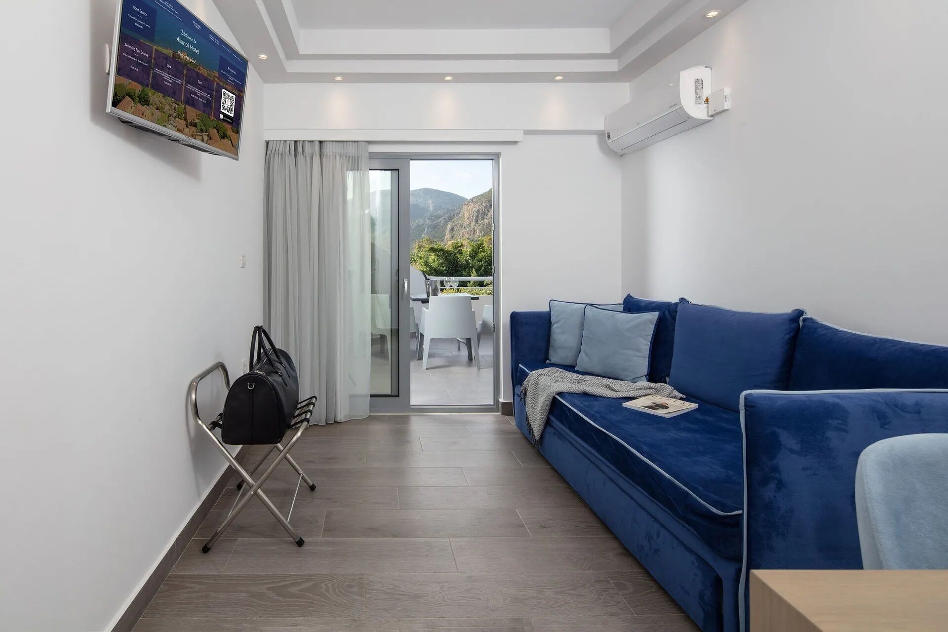 A modern hotel room with a blue sofa, a small table, and a view of the outdoors through a glass door.