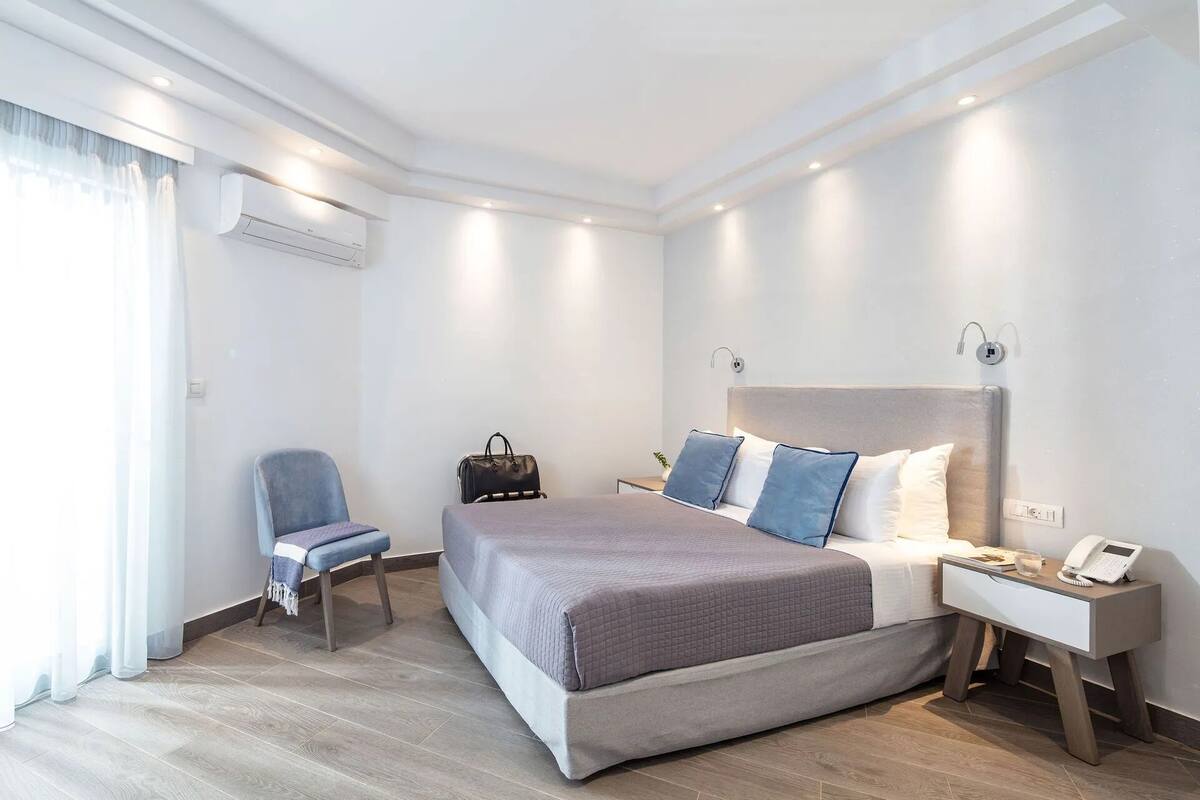 A modern hotel room with a large bed, a chair, a bedside table with a phone, and an air conditioning unit.
