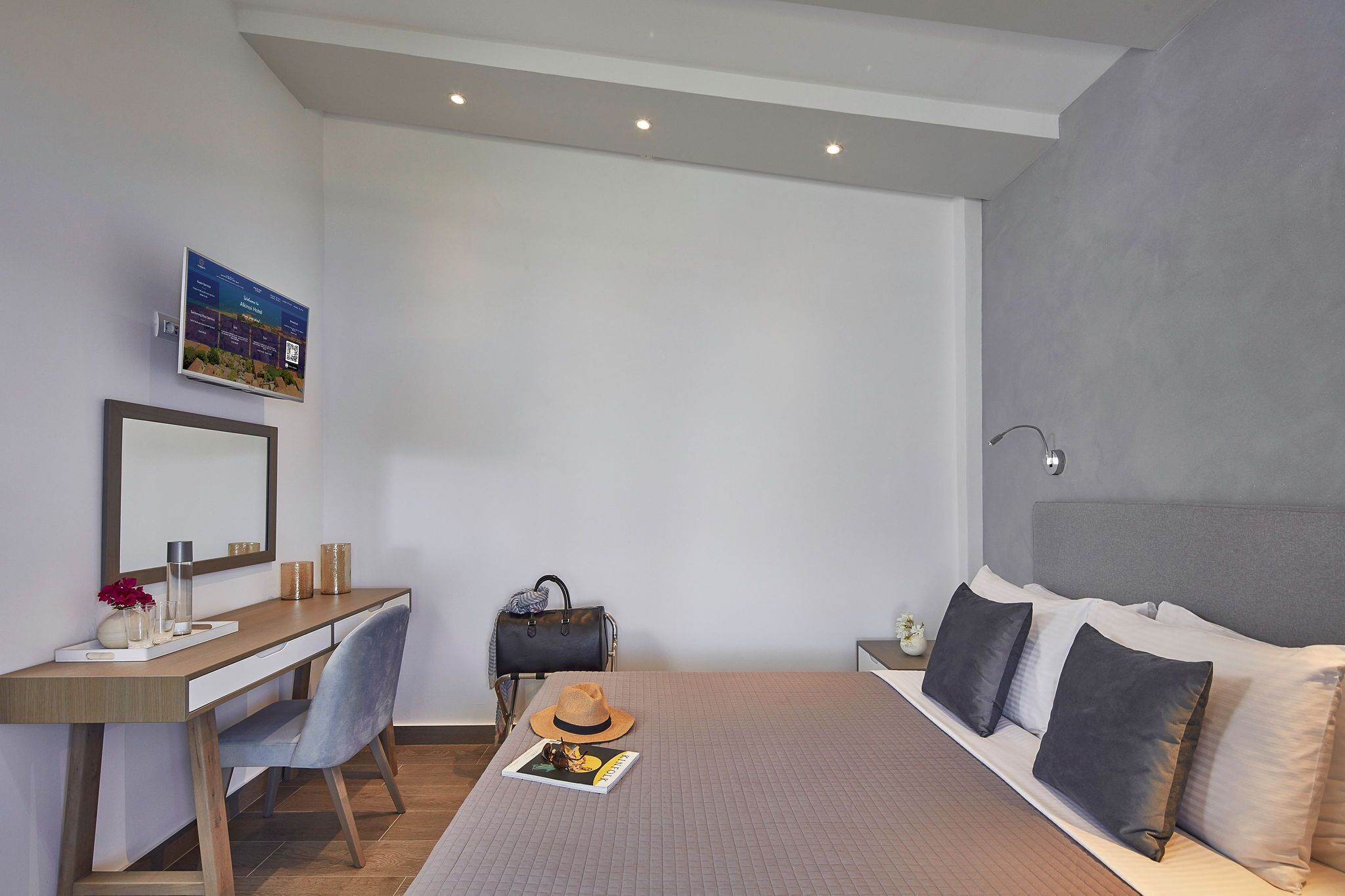 A modern hotel room with a bed, a desk, a chair, a handbag, a hat, and a framed picture on the wall.