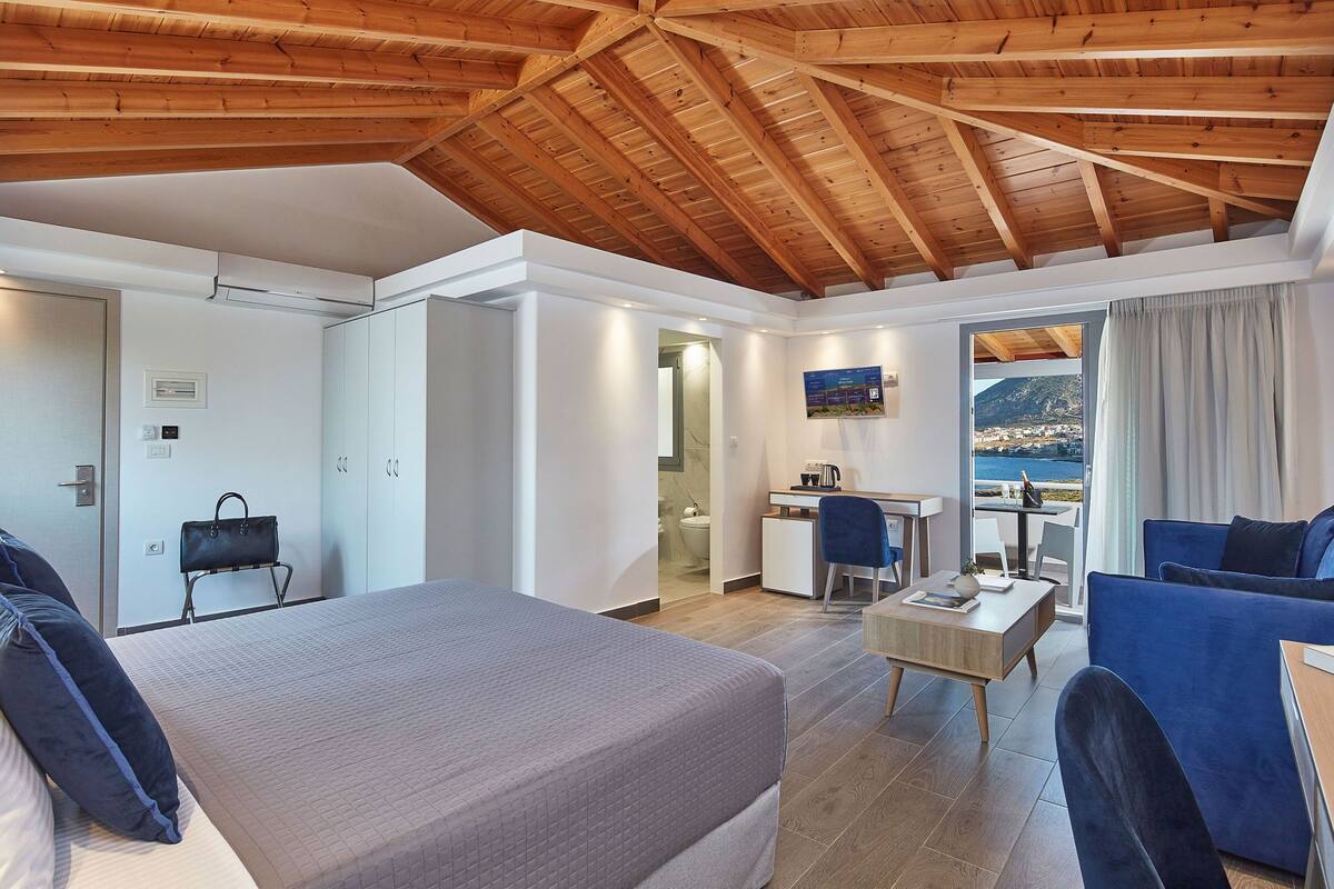 A hotel room with a wooden ceiling, a bed, a desk, a chair, a sofa, and a view of the sea.