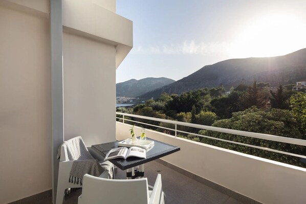 A balcony with a table, a chair, and a book. The balcony overlooks a mountainous landscape with greenery and a clear sky.