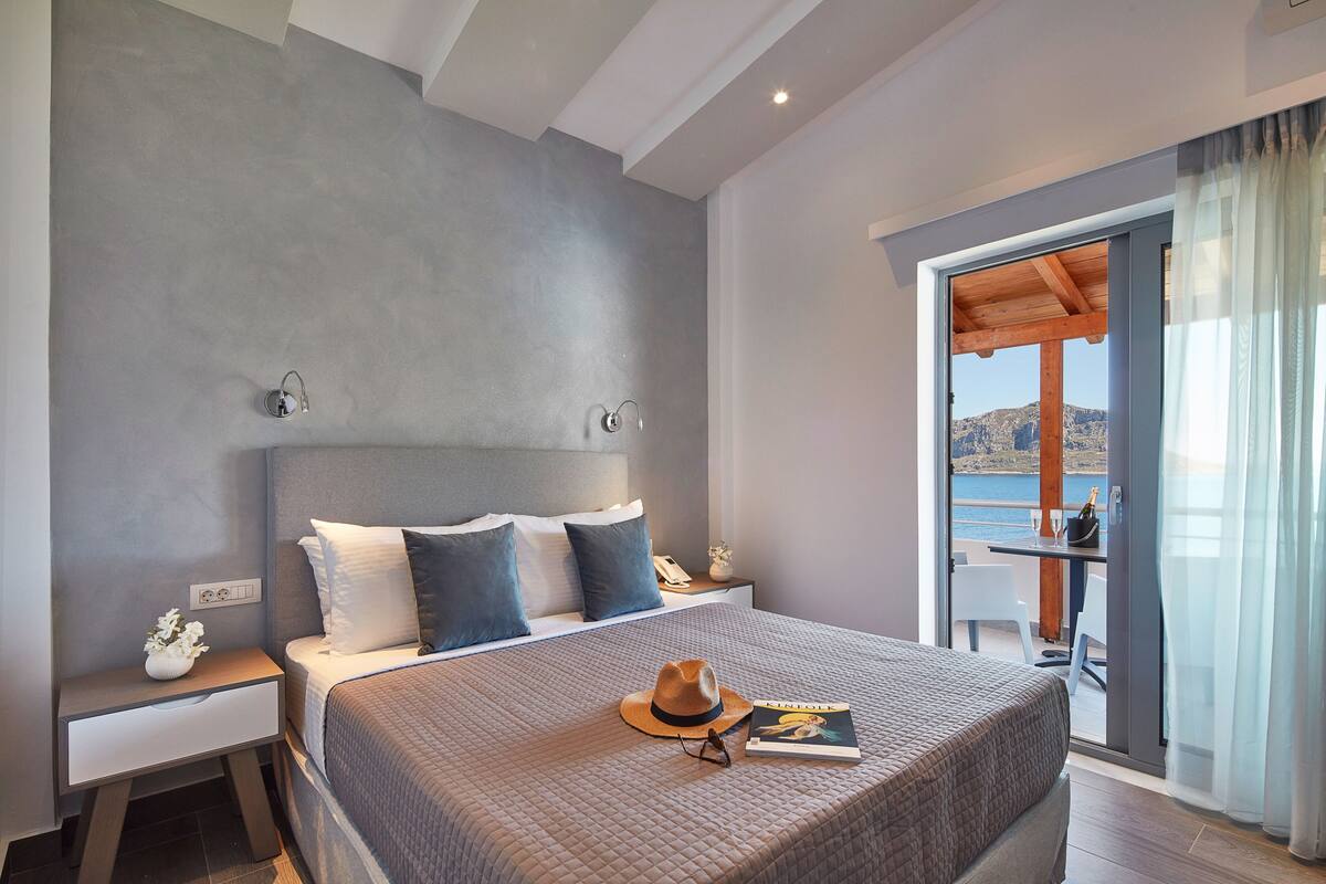 A modern hotel room with a large bed, a nightstand with a phone and vase, and a balcony with a table and chairs.