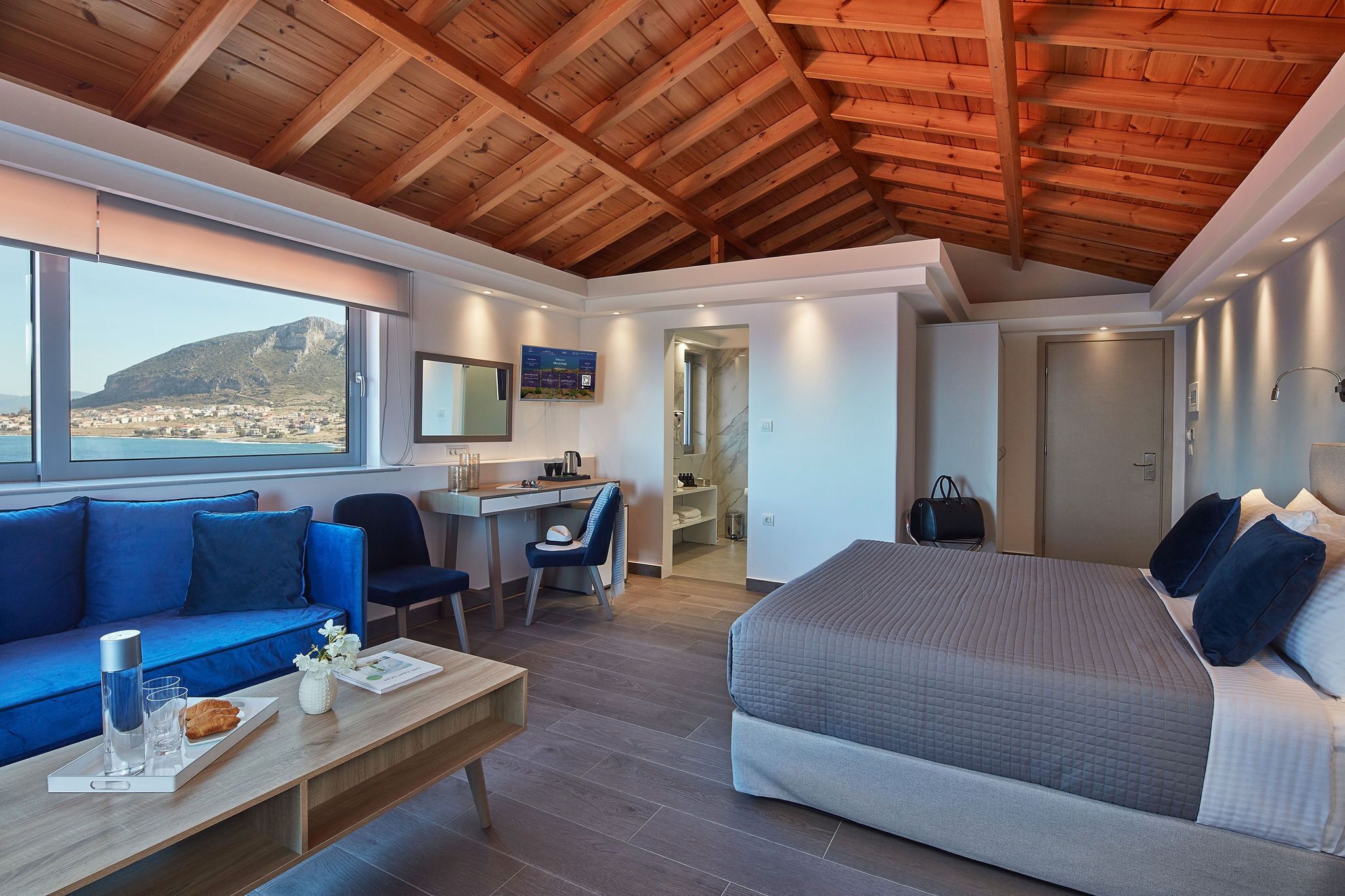 Una habitación de hotel moderna con techo de madera, una cama grande, un sofá azul, un escritorio y vistas al mar.