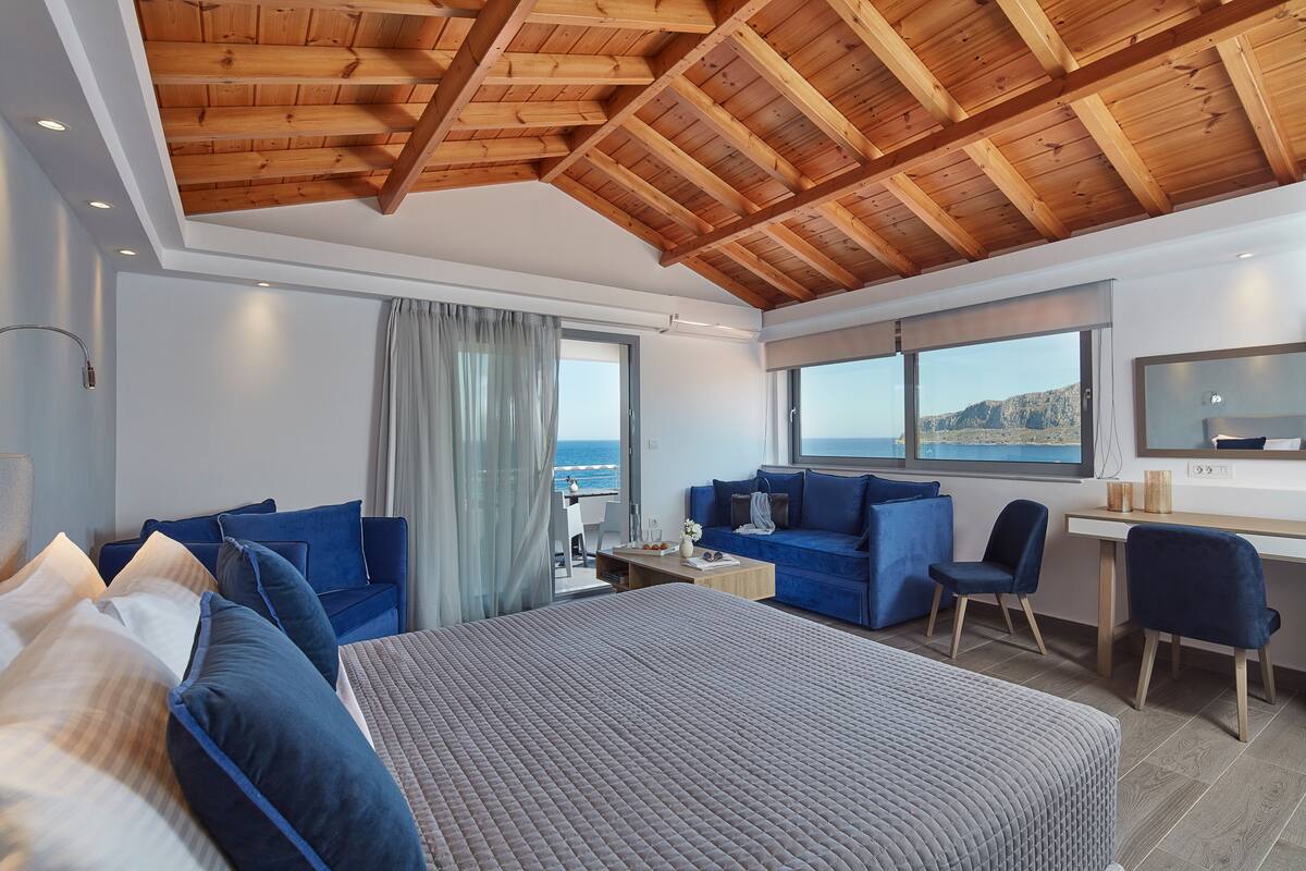 A modern hotel room with a large bed, a sofa, a desk, and a view of the sea.