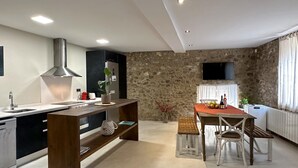 A modern kitchen with a central island, a dining area with a table and chairs, a stone wall, and a mounted television. - Rural House La Païssa (Ullà)
