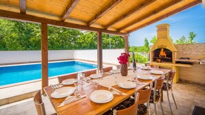 Ein Essbereich im Freien mit Pool, einer Holzpergola und einem Backofen aus Ziegeln.