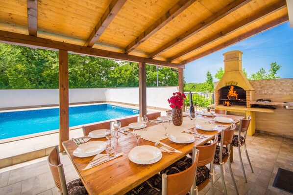 An outdoor dining area with a pool, wooden pergola, and a brick oven.