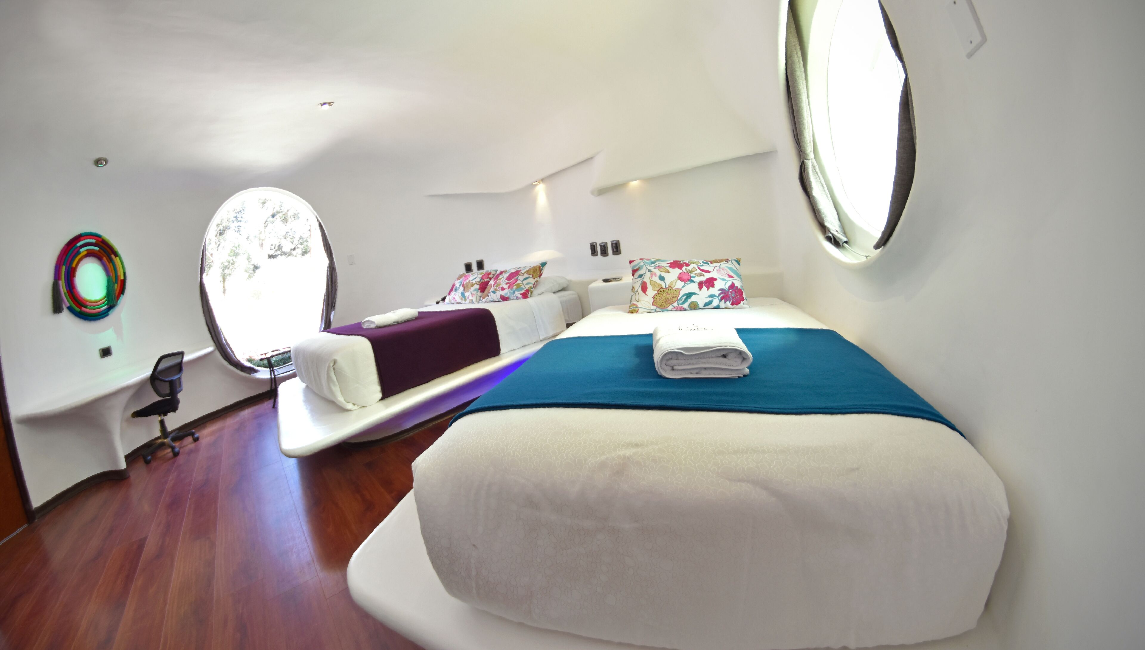 Un dormitorio moderno con una cama grande, un escritorio y una silla. Hay dos ventanas y un espejo en la pared.