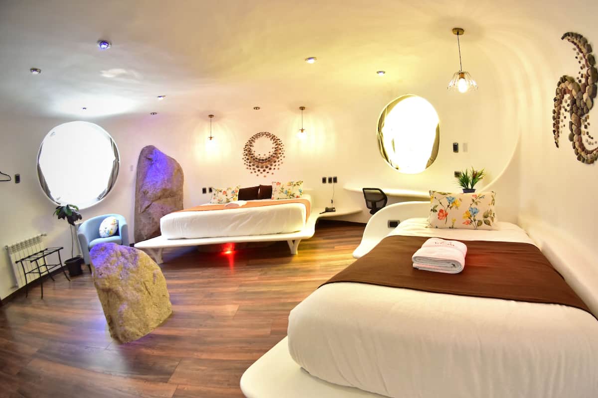 Quarto de hotel moderno com duas camas, um espelho grande, uma janela redonda e piso de madeira.
