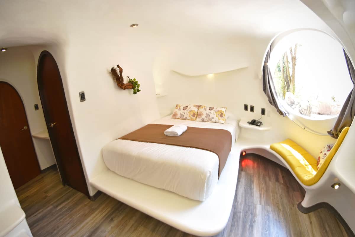 Quarto compacto e moderno, com uma cama, mesa de cabeceira pequena, janela com vista e porta para outro espaço.