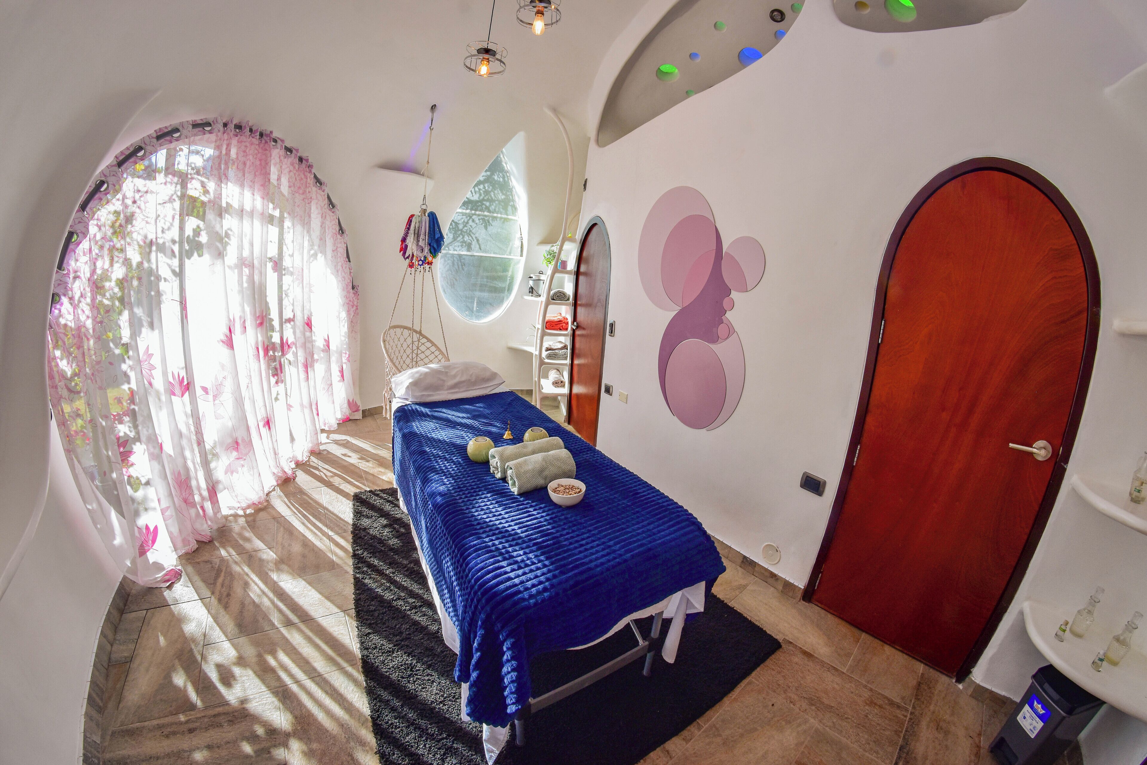 Um quarto com cama, mesa de massagem, janela com cortinas, porta e parede com um desenho circular.