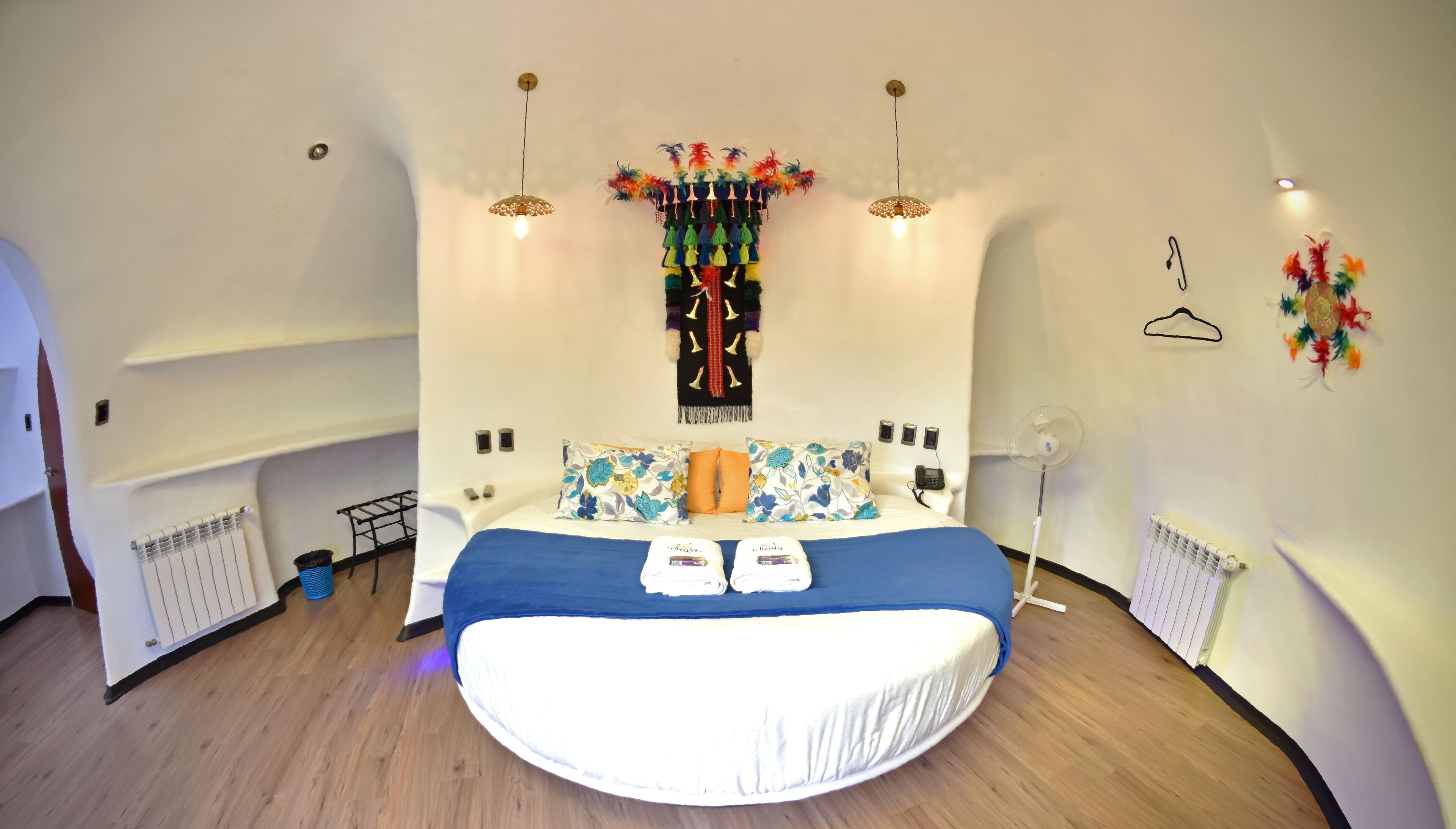A modern bedroom with a curved ceiling, a large bed with blue bedding, a decorative wall hanging, and a standing fan.