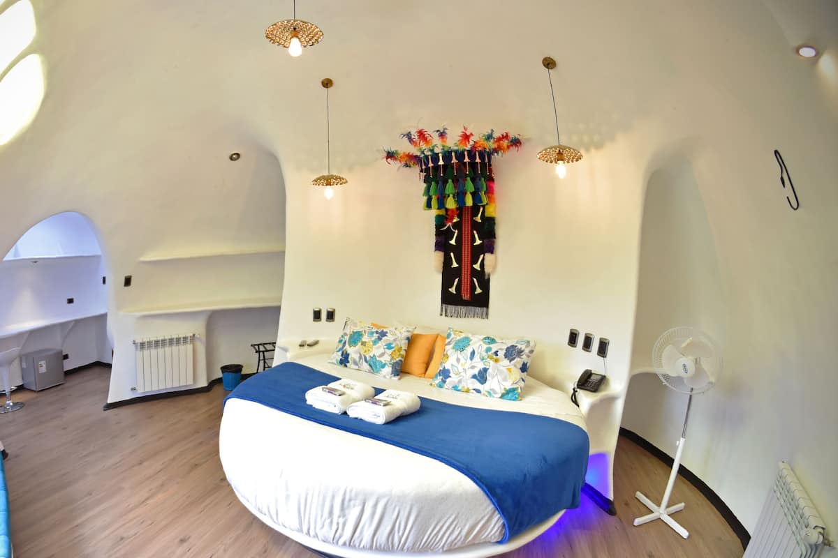 Um quarto moderno com teto curvo, uma cama com colcha azul e branca, um enfeite na parede e um ventilador de chão.