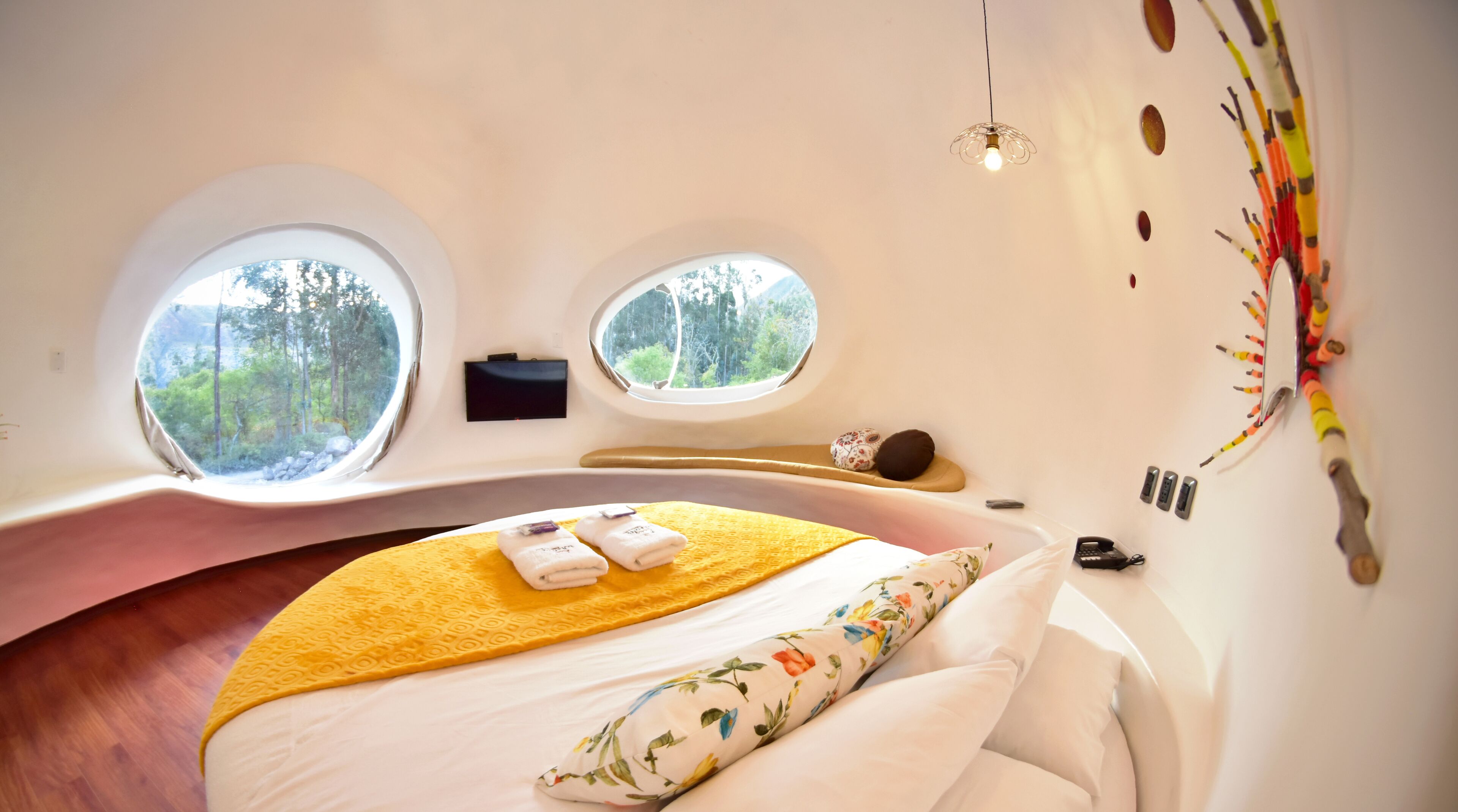 Un dormitorio con techo curvo, dos ventanas con vista al bosque, una cama con manta amarilla y un teléfono sujeto a la pared.