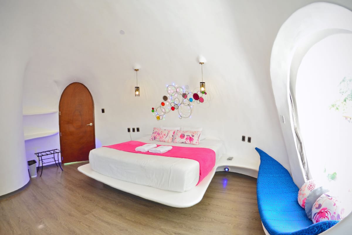 Quarto moderno com teto curvo, cama com colcha rosa e branca, uma cadeira azul e luminária fixada na parede.