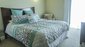 A neatly made bed with patterned bedding, a chair, and a bedside lamp in a room with a large window and light-colored walls.