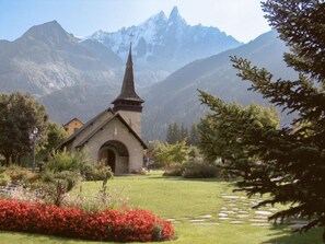 A church with a pointed steeple, surrounded by a well-maintained garden with red flowers, set against a backdrop of mountains. - Champraz by Interhome (Chamonix - Les Praz)