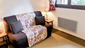 Ein gemütliches Wohnzimmer mit einer Couch, die mit einer gemusterten Decke bedeckt ist, einem kleinen Tisch mit einer Lampe und einem Fenster mit Vorhängen.