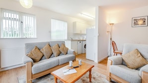 Een moderne woonkamer met een grijze bank, een houten salontafel en een grijze fauteuil. Achtergrond is een keuken zichtbaar met witte kasten en apparatuur.