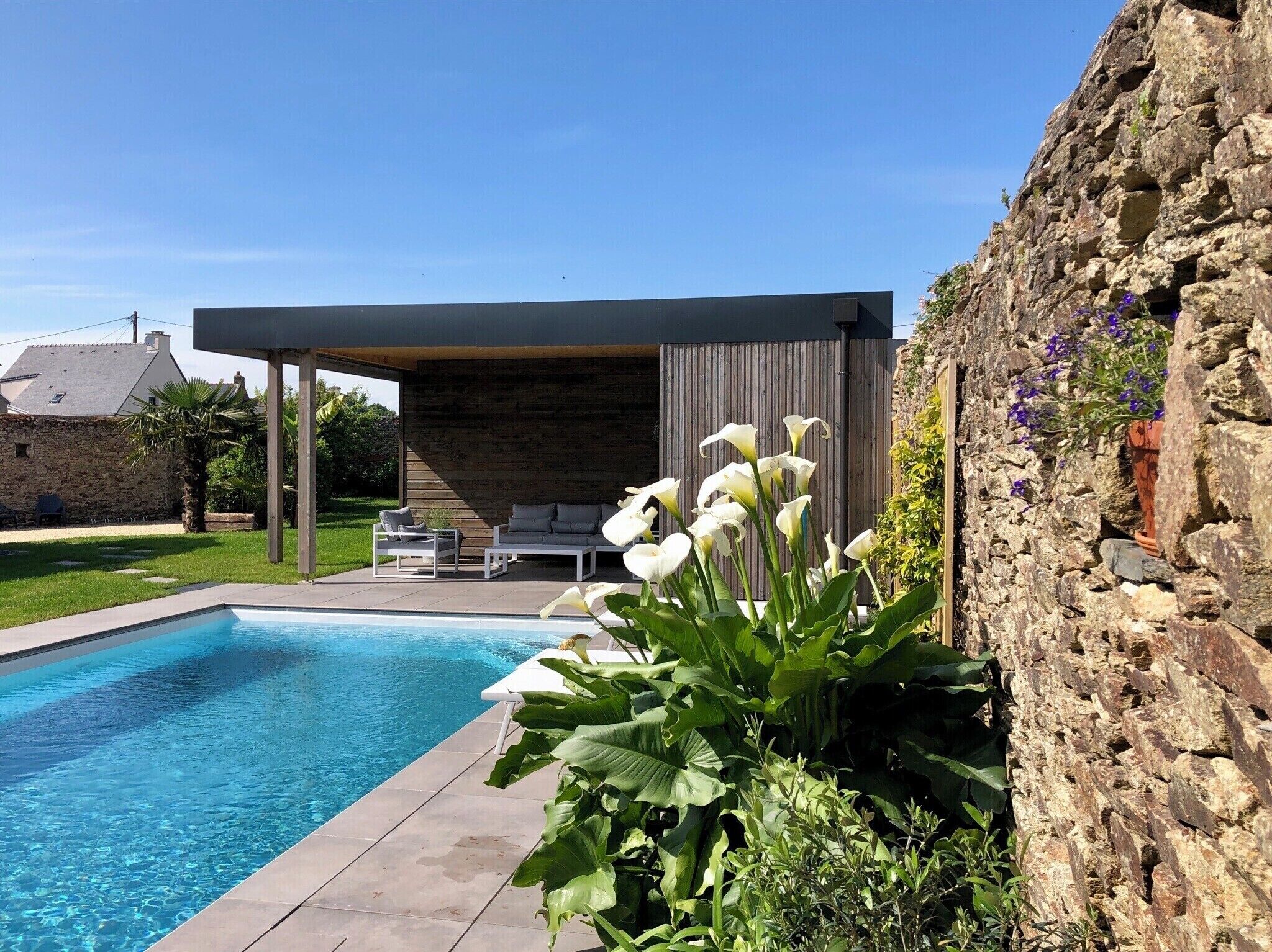 A modern outdoor pool area with a stone wall, a covered seating area, and a variety of plants.