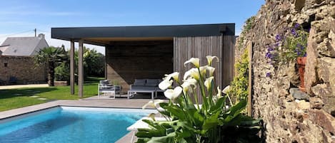 A modern outdoor pool area with a stone wall, a covered seating area, and a variety of plants.