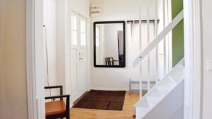 Un couloir avec un plancher en bois, un paillasson marron, une chaise, une porte blanche et un miroir à cadre noir accroché au mur.