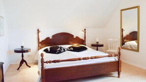 Une chambre à coucher avec un lit à baldaquin en bois, une table d’appoint ronde, une petite table avec une bougie, une chaise blanche et un grand miroir au mur.