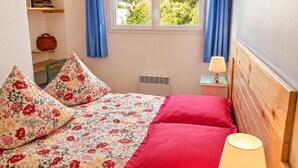A bedroom with a bed, a nightstand, a lamp, a window with curtains, and a shelf with books and a plant. - The waves of money - Le Lavandou (LE LAVANDOU)