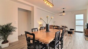 Un coin repas avec une table en bois, des chaises noires et un vase rempli de fleurs blanches. On aperçoit une plante en pot sur la gauche et un ventilateur de plafond au-dessus.