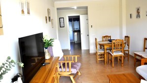 Een woonkamer met een televisie, houten meubels, een eettafel met stoelen en een keuken op de achtergrond.