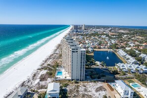 An aerial view of a coastal area with a beach, a tall hotel building, and a swimming pool.