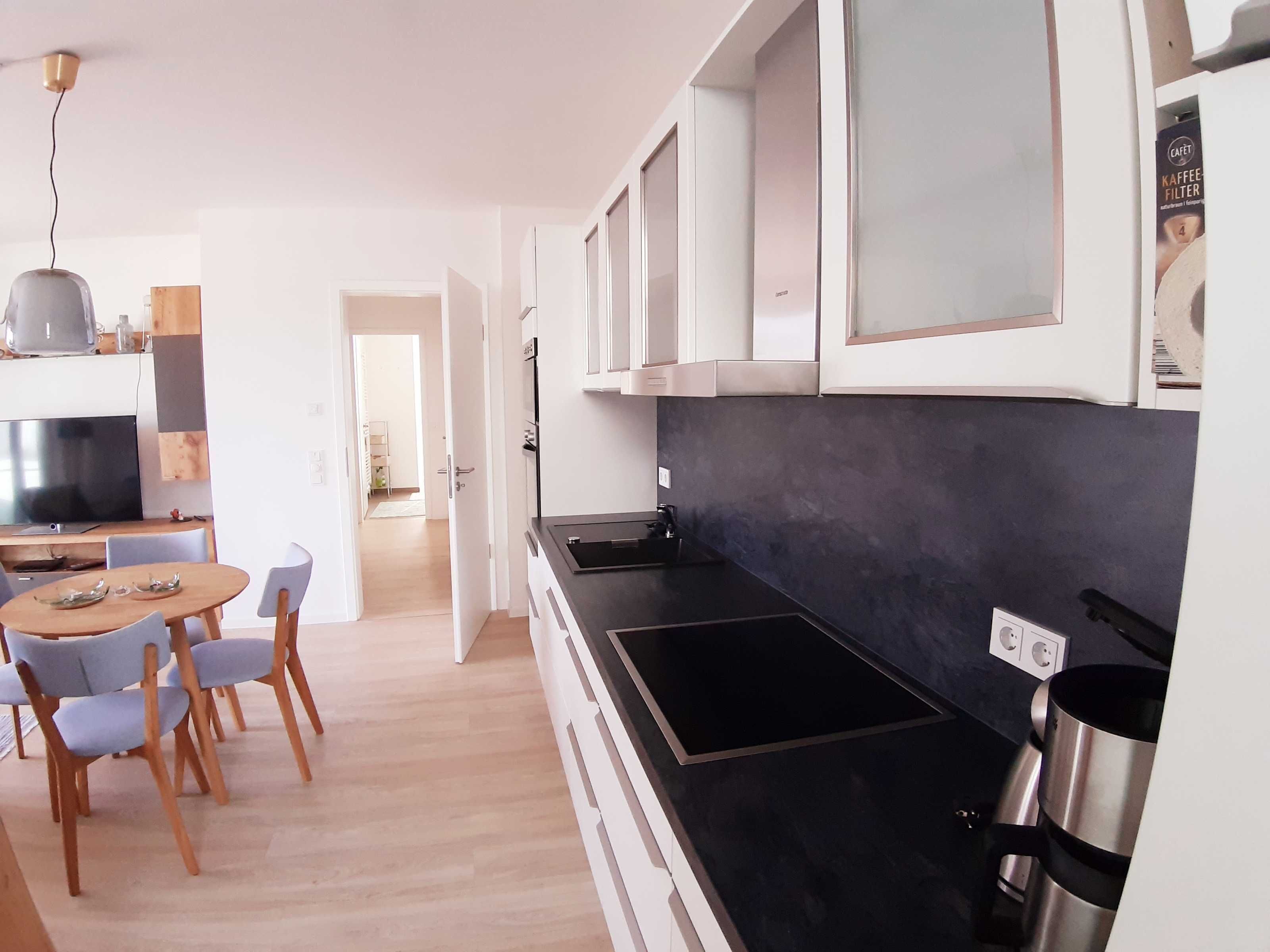 Une cuisine moderne avec des meubles blancs, un plan de travail noir et une bouilloire en acier inoxydable. On y trouve également un coin repas avec une table et des chaises en bois.