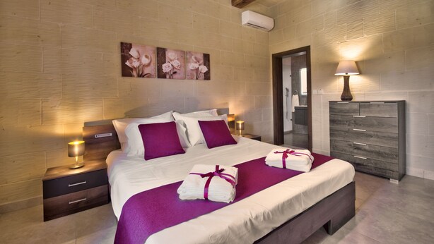 A bedroom with a large bed, purple bedding, a wooden headboard, a nightstand with a lamp, and a dresser with a mirror.