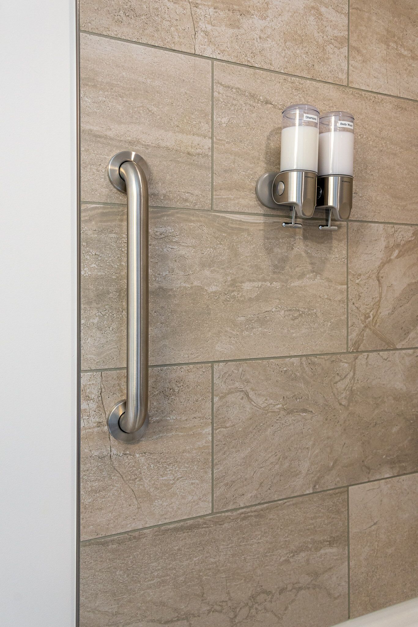 Un baño con paredes de azulejos color beige, una barra de apoyo de acero inoxidable y dos dispensadores de jabón.