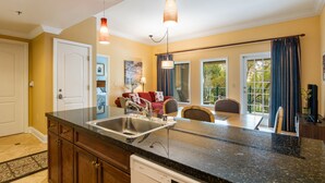 A kitchen with a granite countertop, stainless steel sink, and a dishwasher. There is a dining area with a table and chairs, and a view of the outdoors through large windows. - Gorgeous condo, super comfy, with view of the pool in Lake Las Vegas Village. (Henderson)