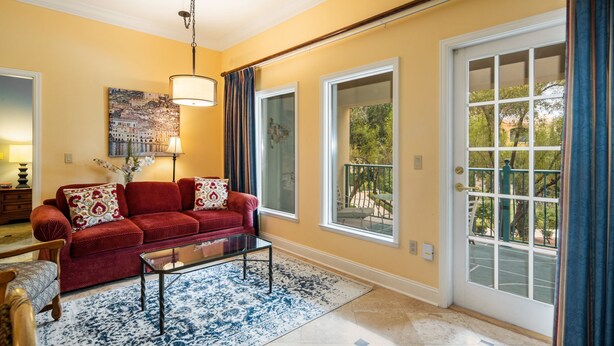 A living room with a red sofa, a glass coffee table, and a blue patterned rug. There are two large windows with curtains, a painting on the wall, and a lamp. - Gorgeous condo, super comfy, with view of the pool in Lake Las Vegas Village. (Henderson)