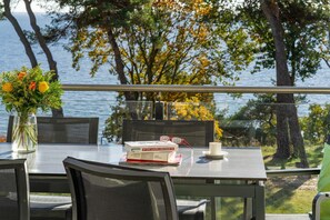 Ein Tisch mit einem Buch, Brillen und einer Blumenvase, mit Blick auf ein Gewässer und Bäume.
