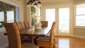 Un espace repas avec une table en bois et des chaises en osier, de grandes fenêtres et une vue sur l’extérieur.