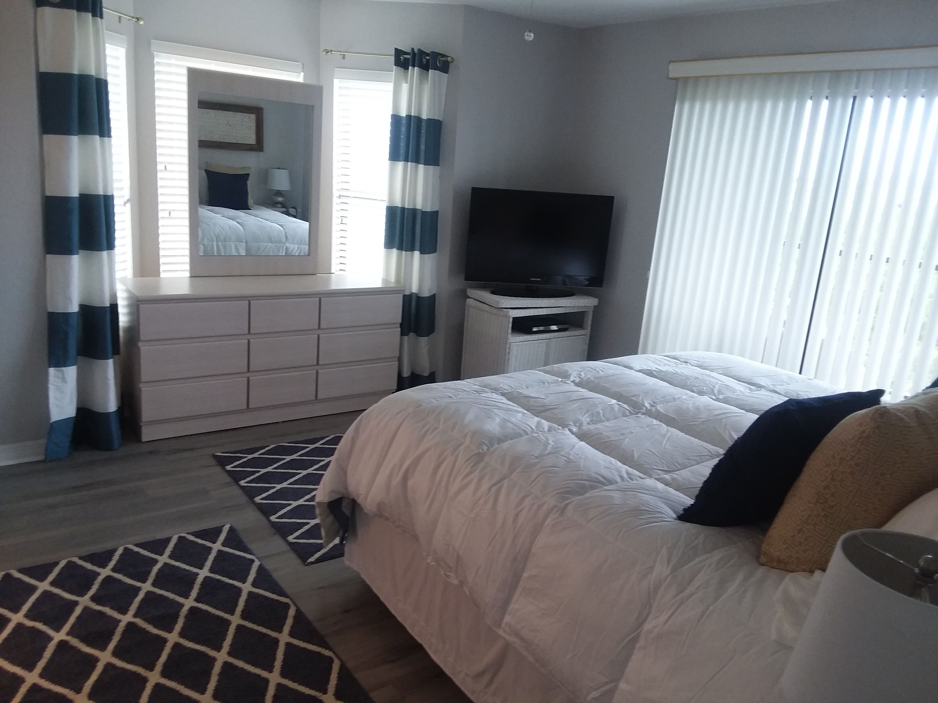Un dormitorio con cama, televisor, cómoda y ventana con cortinas.