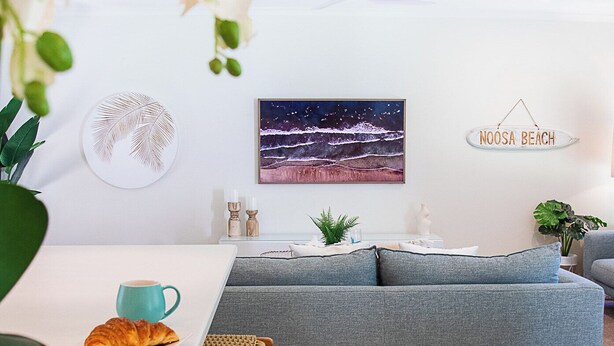 A modern living room with a grey sofa, a dining area with wicker chairs, and a wall-mounted sign reading "NOOSA BEACH". - Noosa Heads Endless Summer Beach Retreat (Noosa Heads)