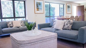 A living room with two grey sofas, a white woven ottoman, and a dining area with a table and chairs. - Noosa Heads Endless Summer Beach Retreat (Noosa Heads)