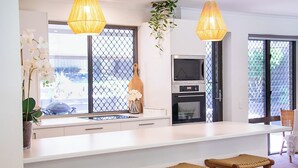 A modern kitchen with a white countertop, two hanging pendant lights, a built-in oven, and a TV. There are three wicker stools at the counter. - Noosa Heads Endless Summer Beach Retreat (Noosa Heads)