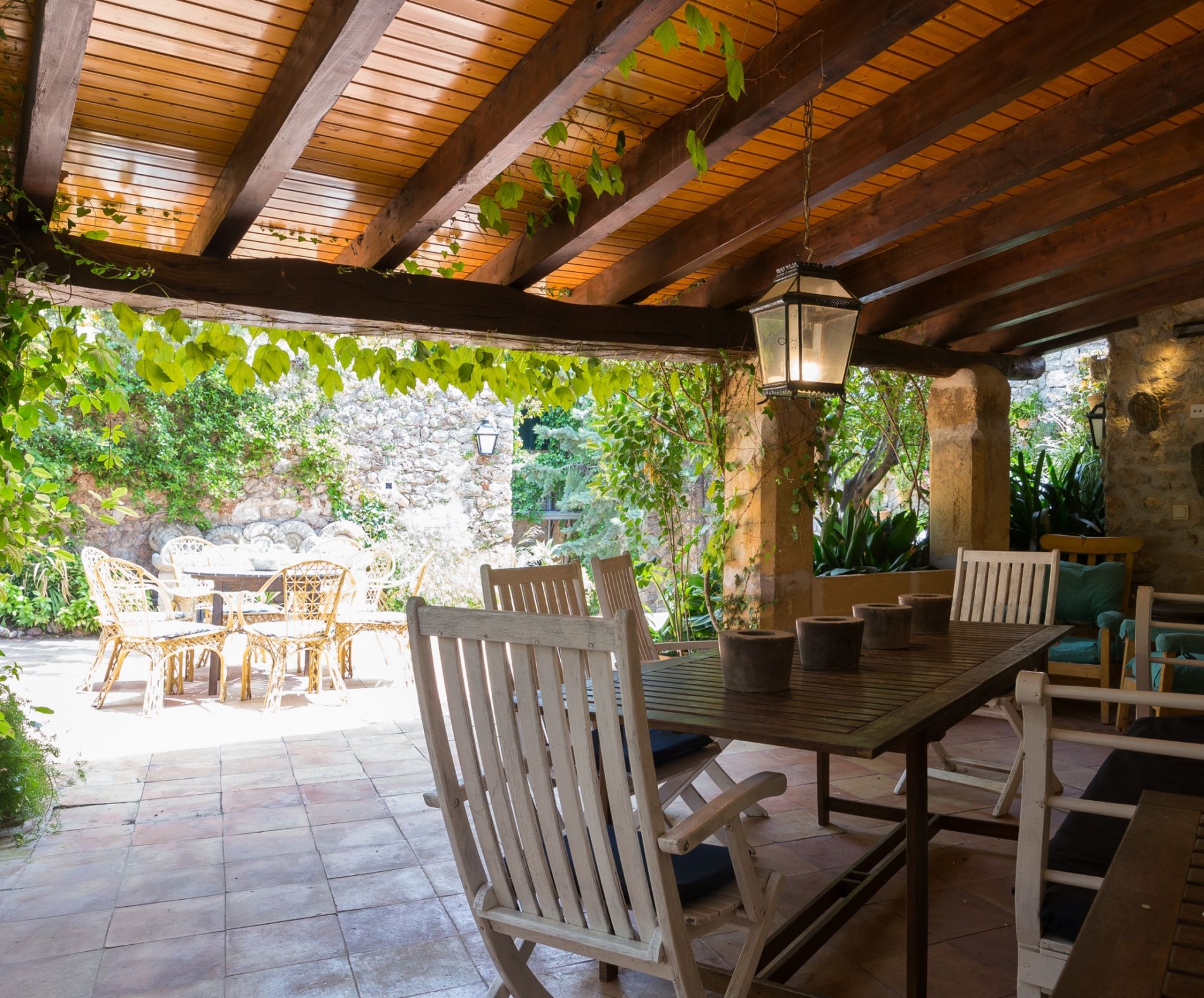 An outdoor patio with wooden tables and chairs, stone walls, and hanging plants.
