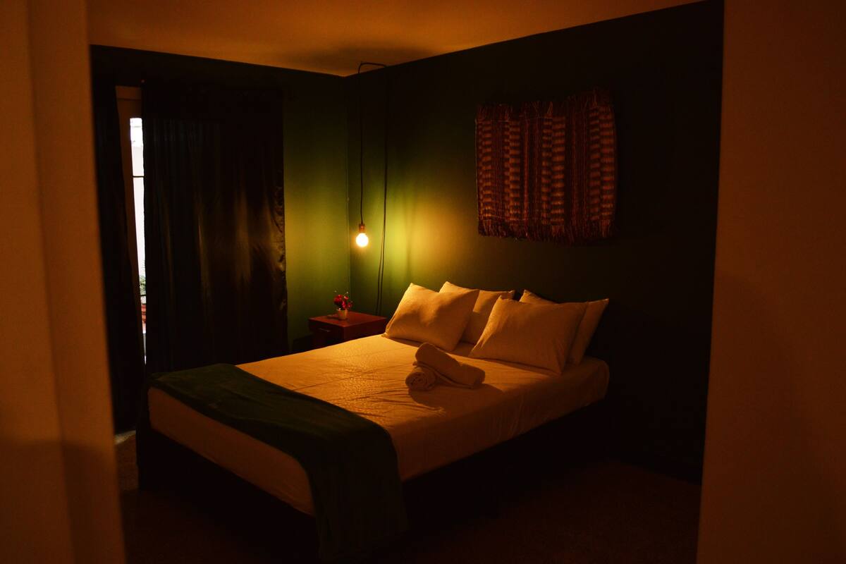 A dimly lit hotel room with a bed, a nightstand, and a wall-mounted lamp.