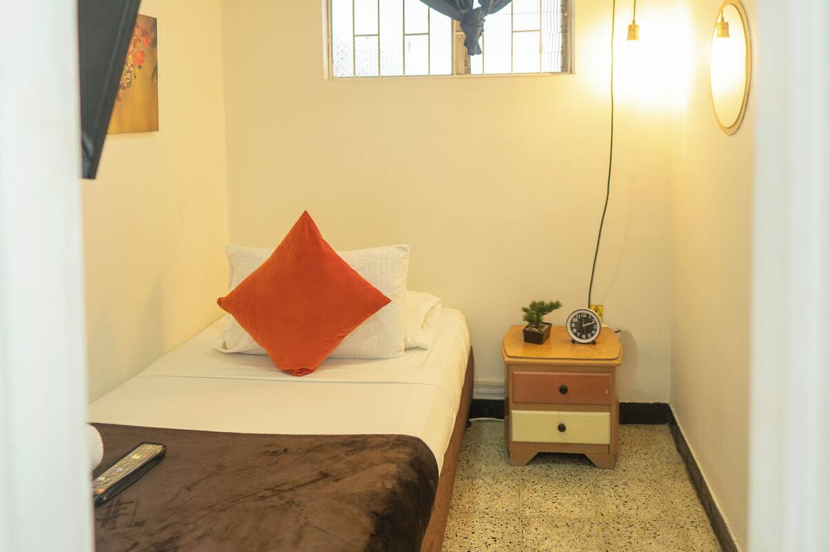 A bedroom with a bed, a nightstand with a clock and a plant, a window with bars, and a wall-mounted light fixture.