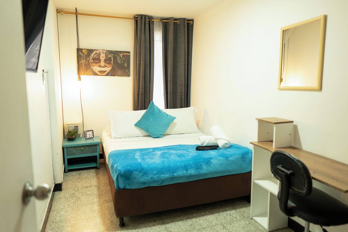 A bedroom with a bed, a desk, a chair, a nightstand, a lamp, and a painting on the wall.