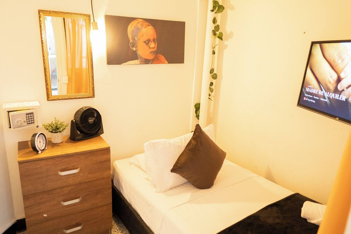 A hotel room with a bed, a bedside table with a clock and a plant, a dresser with drawers, a mirror, and a TV screen displaying an image.