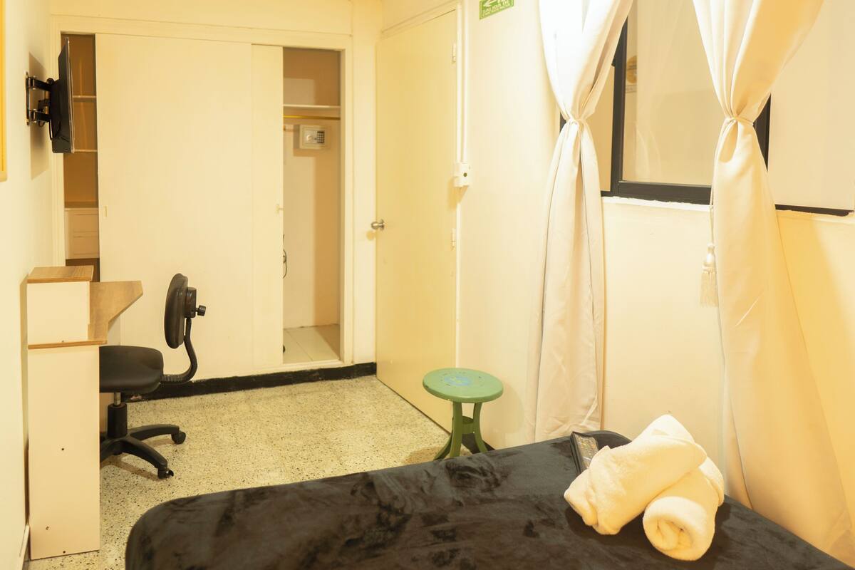 A hotel room with a bed, a desk with a chair, a small stool, a TV mounted on the wall, and a window with curtains.
