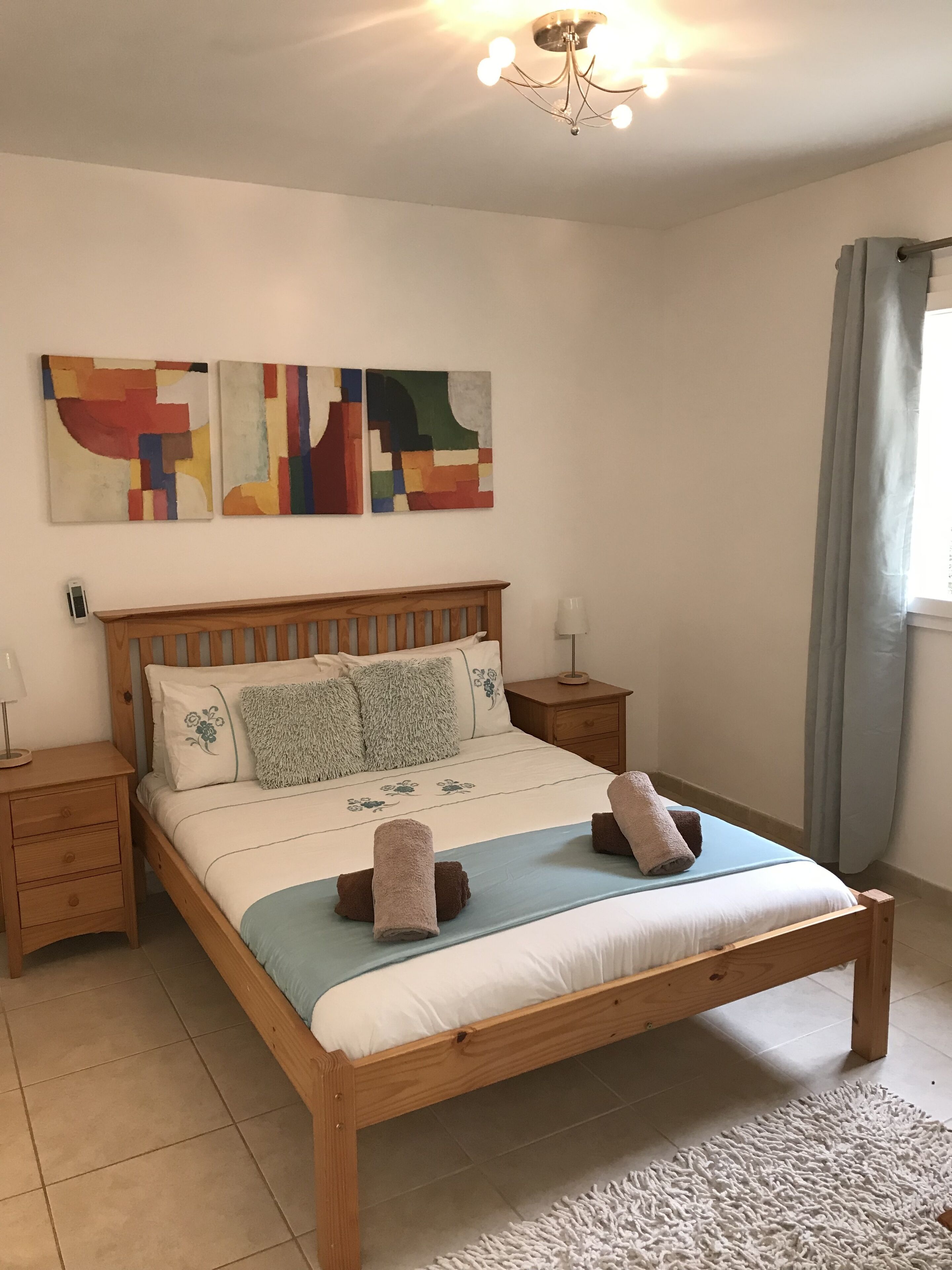 A bedroom with a wooden bed, two bedside tables, a nightstand, a wall-mounted light fixture, and abstract artwork.