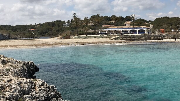 A coastal view with a beach, clear blue waters, and a building with a blue roof and white walls. - Luxurious villa near beach: swimming pool (heatable), whirlpool, high speed WiFi (Cala Murada)